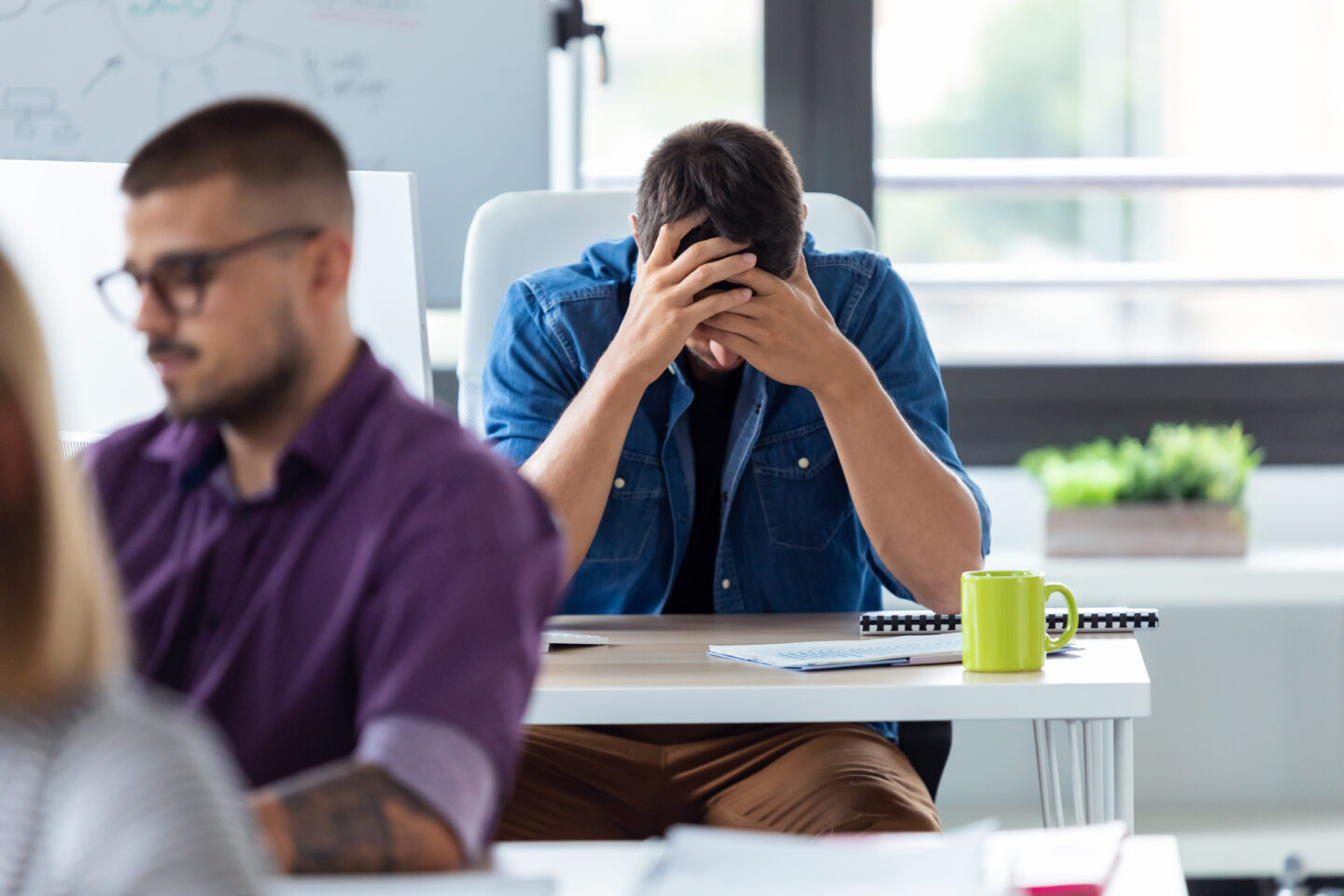 young businessman holding his face with hands while sitting at the desk in the creative office