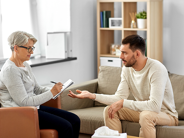 Male sat on sofa during therapy session with an older woman with glasses taking notes