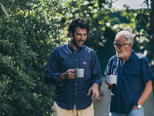 A dark haired man strolling outdoors alongside an older man with glasses, both holding mugs