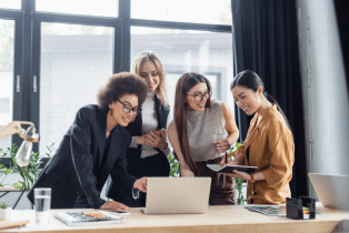 A group of female employees huddled around a laptop and smiling in an office environment