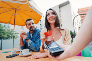 Woman holding a credit card above a card reader smiling whilst sat next to a male in an outdoor cafe setting