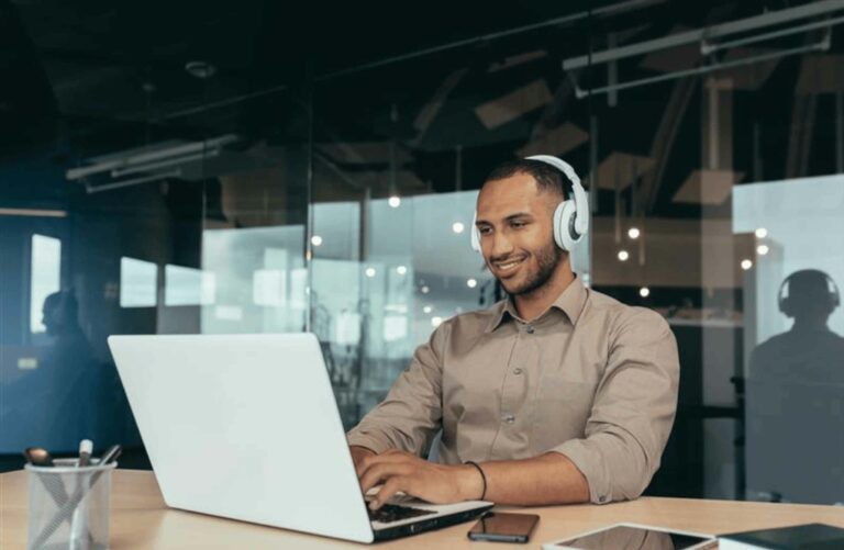 Smiling man with headphones on sat at a desk looking at a laptop